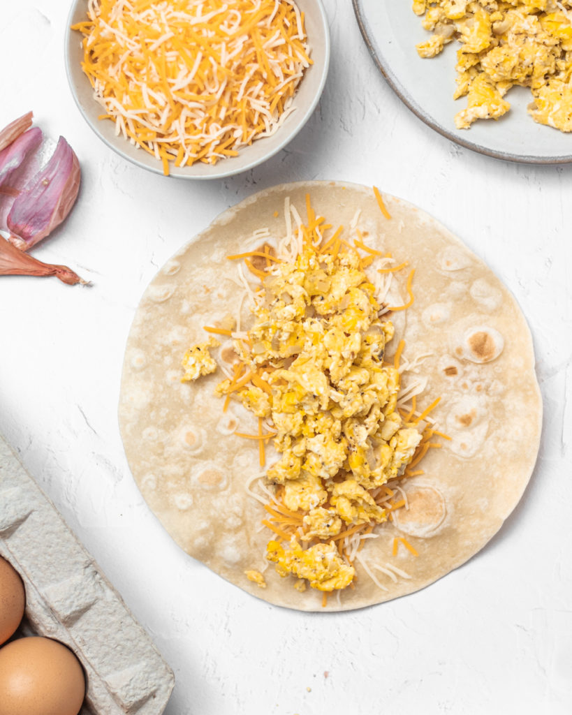 breakfast enchiladas in the making; scrambled eggs and cheese on a flat tortilla ready to be rolled